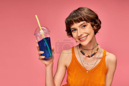 Téléchargez les photos : Positive jeune femme aux cheveux bruns courts tenant boisson rafraîchissante d'été sur fond rose - en image libre de droit