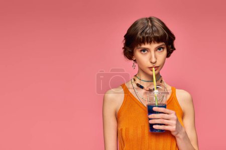 junge Frau mit kurzen brünetten Haaren trinkt blauen erfrischenden Sommercocktail auf rosa Hintergrund