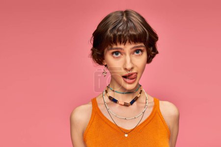 cheeky playful girl in 20s with short brunette hair sticking tongue out on pink background