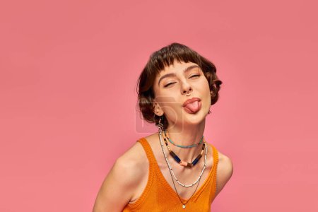 cheeky carefree girl in 20s with short brunette hair sticking tongue out on pink background