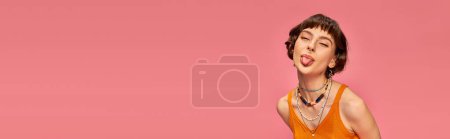 cheeky playful girl in 20s with short brunette hair sticking tongue out on pink background, banner