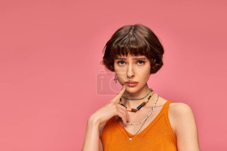 worried young woman in her 20s standing in orange knitted tank top on pink background, concern