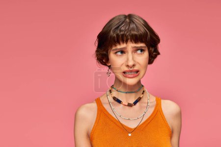 puzzled young girl in her 20s standing in orange knitted tank top on pink background, unsure