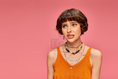 puzzled young woman in her 20s standing in orange knitted tank top on pink background, uneasy