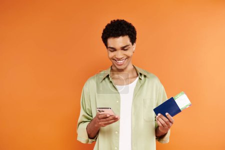Téléchargez les photos : Séduisant homme afro-américain joyeux en tenue confortable regardant le téléphone et tenant passeport et billet - en image libre de droit