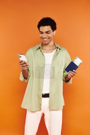 Téléchargez les photos : Beau Africain Américain joyeux en tenue confortable regardant le téléphone et tenant passeport et billet - en image libre de droit