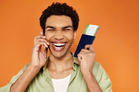 Téléchargez les photos : Joyeux homme afro-américain attrayant en tenue confortable parlant par téléphone tenant passeport avec billet - en image libre de droit