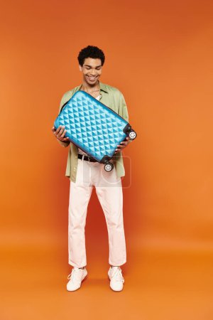 Photo for Cheerful african american man in casual outfit looking at blue suitcase in hands on orange backdrop - Royalty Free Image