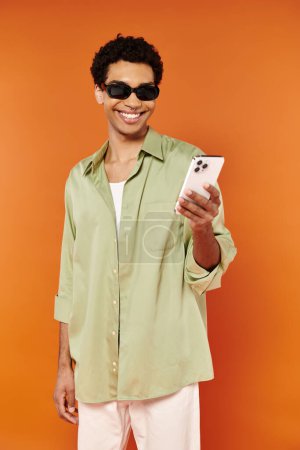 Photo for Good looking jolly african american man with trendy sunglasses looking at phone on orange backdrop - Royalty Free Image