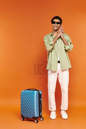 Photo for Appealing happy african american man with trendy sunglasses talking by phone near blue suitcase - Royalty Free Image