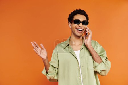 Photo for Handsome merry african american man with stylish sunglasses talking by phone on orange backdrop - Royalty Free Image