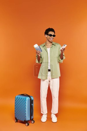 Téléchargez les photos : Joyeux homme afro-américain avec des lunettes de soleil tenant billet et passeport à côté de sa valise bleue - en image libre de droit