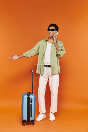 Photo for Joyous african american man with trendy sunglasses talking by phone next to is blue suitcase - Royalty Free Image