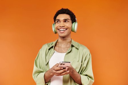 Téléchargez les photos : Élégant homme afro-américain joyeux avec écouteurs tenant le téléphone et détournant les yeux, fond orange - en image libre de droit