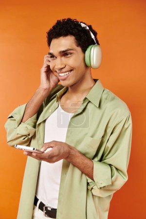 Téléchargez les photos : Homme afro-américain joyeux à la mode avec écouteurs tenant le téléphone et détournant les yeux, fond orange - en image libre de droit