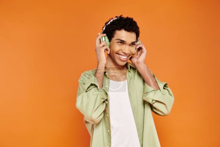 Téléchargez les photos : Joyeux homme afro-américain élégant en tenue confortable avec écouteurs détournant les yeux, toile de fond orange - en image libre de droit