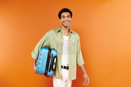 Téléchargez les photos : Joyeux jeune homme afro-américain en vêtements décontractés tenant sa valise et souriant à la caméra - en image libre de droit