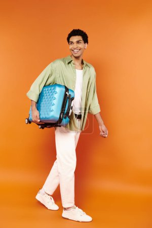 Téléchargez les photos : Beau Africain américain joyeux en tenue confortable marchant avec sa valise bleue à la main - en image libre de droit