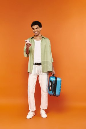 Photo for Joyful young african american man looking at his mobile phone and holding suitcase, orange backdrop - Royalty Free Image