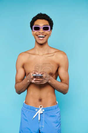 Téléchargez les photos : Joyeux homme afro-américain avec des lunettes de soleil élégantes dans les maillots de bain tenant son smartphone - en image libre de droit
