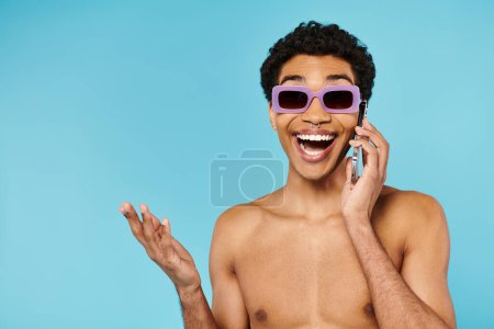 heureux beau homme afro-américain dans les maillots de bain avec des lunettes de soleil élégantes parler par téléphone