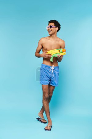 joyous young african american man in swimming trunks with stylish sunglasses posing with water gun