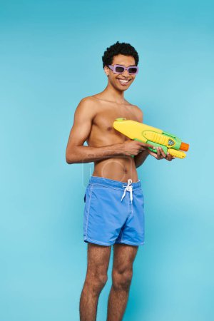 Téléchargez les photos : Joyeux jeune homme afro-américain dans des maillots de bain avec des lunettes de soleil élégantes posant avec pistolet à eau - en image libre de droit