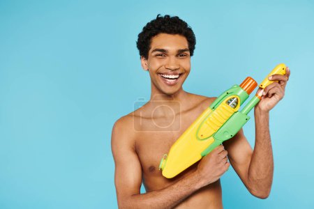positiv ansprechender afrikanisch-amerikanischer Mann posiert mit Wasserpistole und lächelt vor blauem Hintergrund in die Kamera