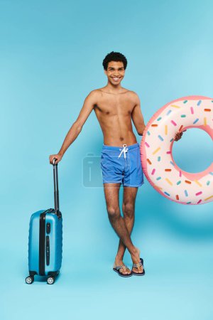 Téléchargez les photos : Homme afro-américain joyeux dans des maillots de bain avec anneau de natation et valise souriant à la caméra - en image libre de droit