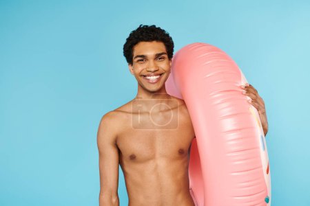 Téléchargez les photos : Homme afro-américain joyeux posant avec anneau de natation sur fond bleu et regardant la caméra - en image libre de droit