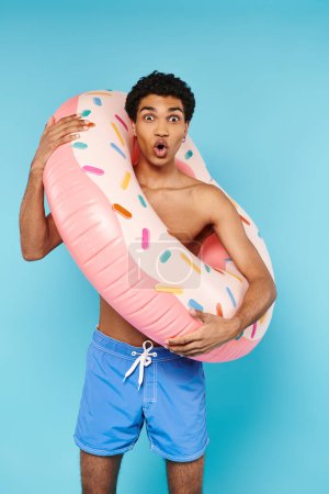 Téléchargez les photos : Homme afro-américain satisfait posant avec anneau de natation sur fond bleu et regardant la caméra - en image libre de droit