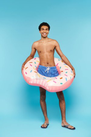 Téléchargez les photos : Attrayant homme afro-américain joyeux avec anneau de natation sur fond bleu en regardant la caméra - en image libre de droit