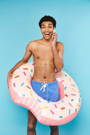 Téléchargez les photos : Beau beau homme afro-américain joyeux dans les troncs de natation avec beignet gonflable parlant par téléphone - en image libre de droit