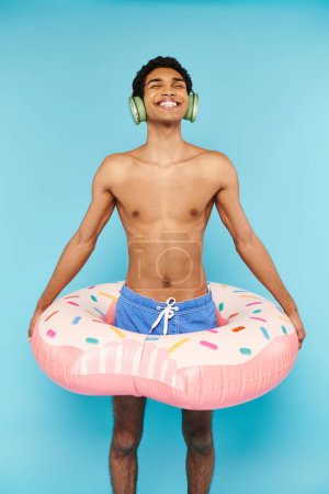 Foto de Hombre americano africano alegre en los baúles de natación con la rosquilla inflable que disfruta de la música en auriculares - Imagen libre de derechos