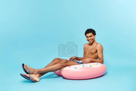 Téléchargez les photos : Homme afro-américain joyeux dans les maillots de bain assis sur beignet gonflable et souriant à la caméra - en image libre de droit