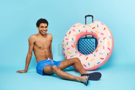 Téléchargez les photos : Positif jeune homme afro-américain dans les maillots de bain assis près de la valise et beignet gonflable - en image libre de droit