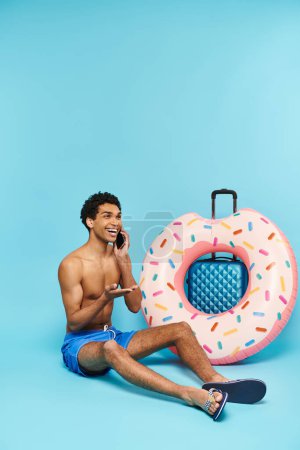 Téléchargez les photos : Joyeux homme afro-américain parlant par téléphone portable à côté de sa valise et beignet gonflable - en image libre de droit