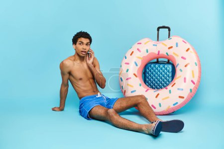 Photo for Young african american man talking by mobile phone next to his suitcase and inflatable donut - Royalty Free Image