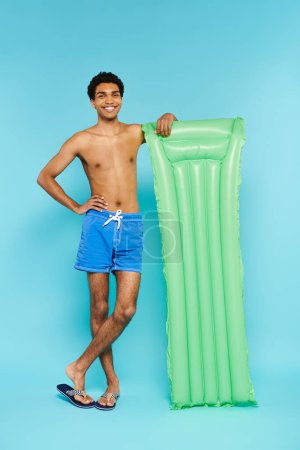 hombre afroamericano positivo en bañador troncos posando con colchón de aire y sonriendo a la cámara