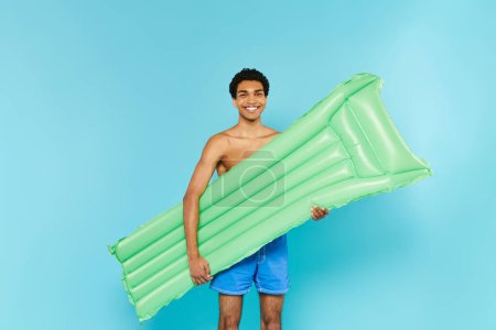 joyous african american man in swimming trunks posing with air mattress and smiling at camera