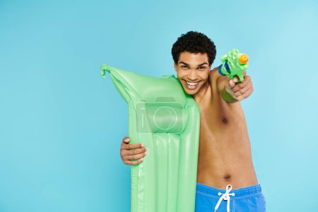 Photo for Cheerful young african american man posing with air mattress and water gun on blue background - Royalty Free Image