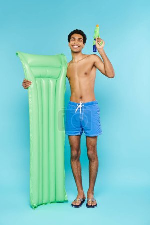 positive young african american man posing with air mattress and water gun on blue background