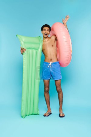 Foto de Alegre afroamericano hombre posando con colchón de aire y donut inflable y sonriendo a la cámara - Imagen libre de derechos