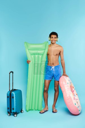 jolly african american man with headphones with air mattress and inflatable donut next to suitcase