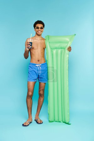merry african american man enjoying cocktail and holding air mattress on blue backdrop, banner