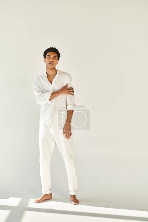 Téléchargez les photos : Élégant homme afro-américain en tenue blanche posant pieds nus et regardant la caméra sur fond beige - en image libre de droit