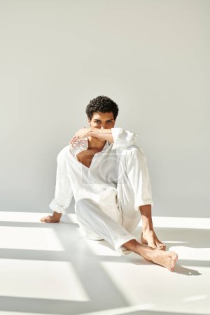 Foto de Atractivo hombre afroamericano sofisticado en traje de lino sentado en el suelo y mirando a la cámara - Imagen libre de derechos