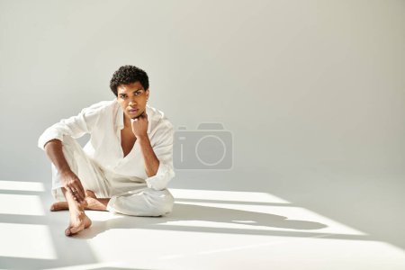 Foto de Refinado joven afroamericano hombre en traje de lino mirando a la cámara en el suelo sobre fondo beige - Imagen libre de derechos
