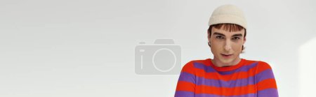 Téléchargez les photos : Homme gay joyeux avec chapeau blanc en tenue vive posant sur fond gris et regardant la caméra, bannière - en image libre de droit
