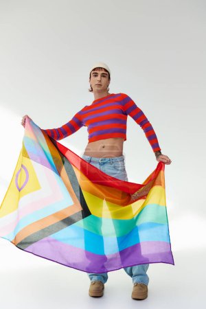 Foto de Alegre atractivo gay hombre en vívido traje con blanco sombrero sosteniendo arco iris bandera y mirando a cámara - Imagen libre de derechos